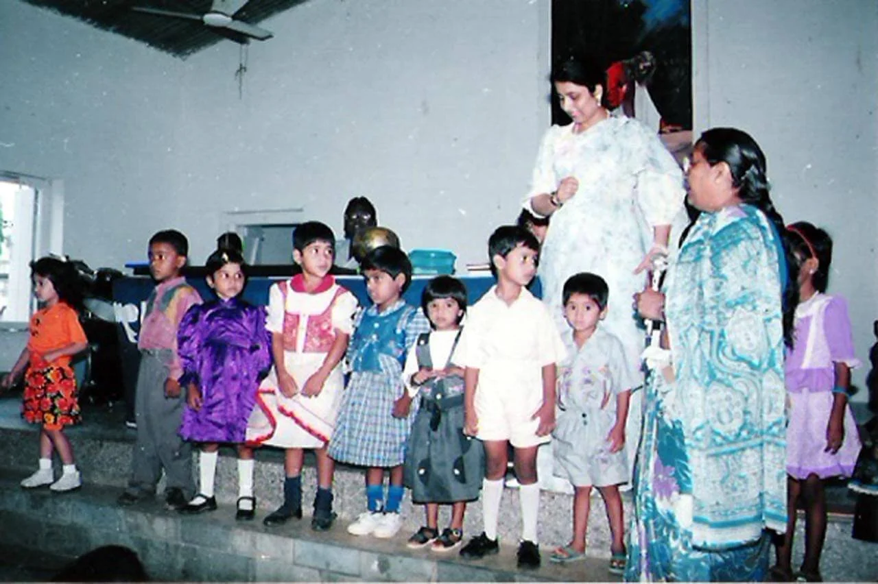 Children standing in a line on a stage with a woman, likely a teacher, overseeing them.
