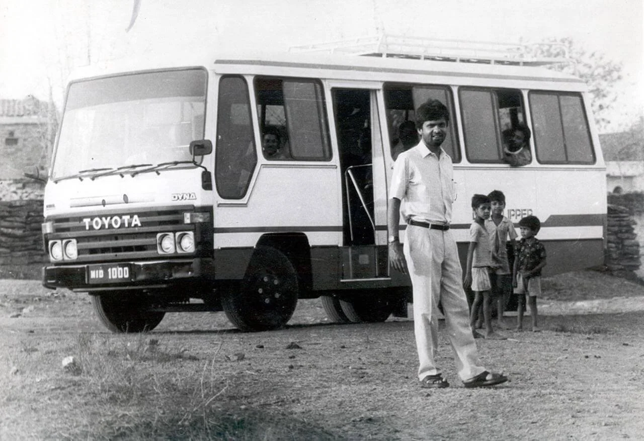 Black and white photo of a man standing in front of a vintage Toyota bus with children inside and outside, on a dirt road