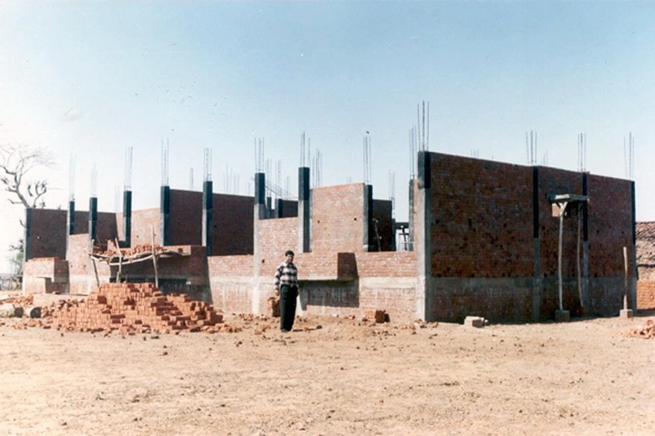 A man standing in front of an under-construction brick building with unfinished walls and vertical reinforcement bars.