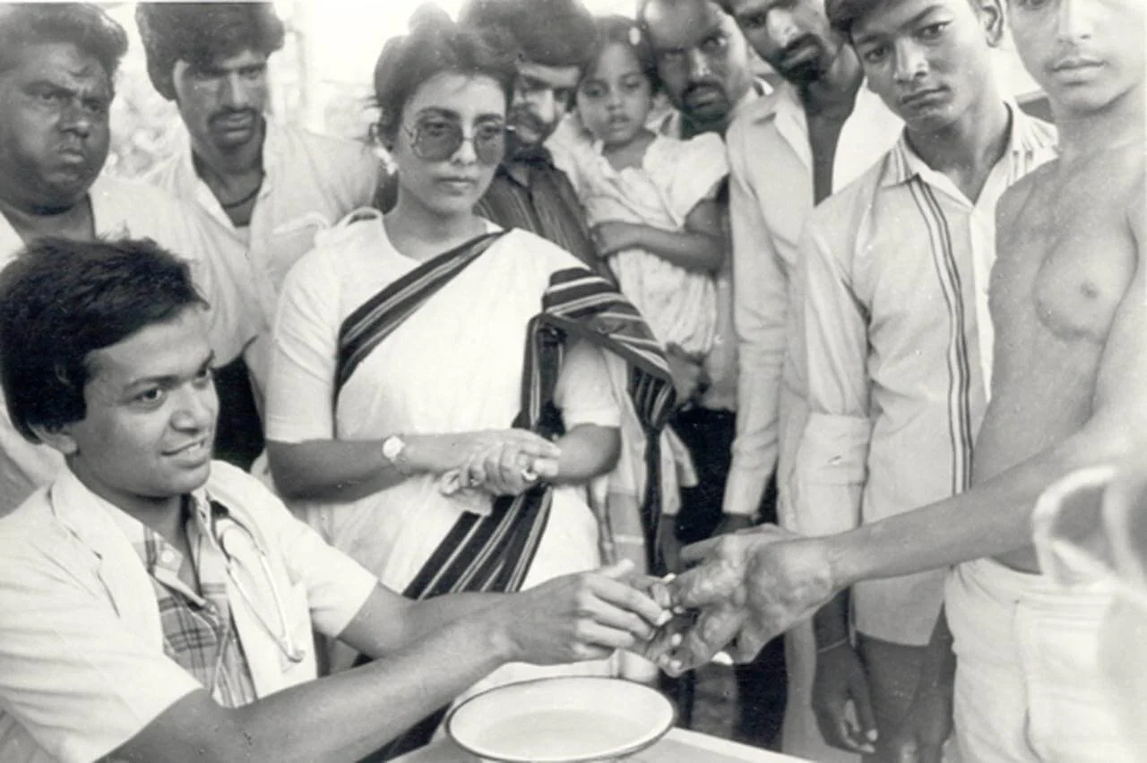 Black and white photograph of a doctor giving an injection to a shirtless man in front of a group of men and women observing.