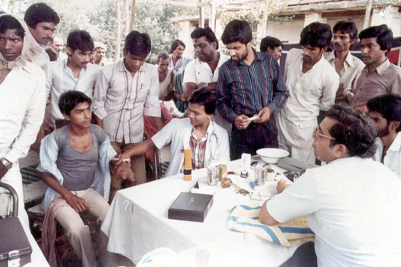 A young man in a medical coat shaking hands with a teenage boy, surrounded by a group of men, some in white, gathered around a table with medical supplies and documents in an outdoor setting.