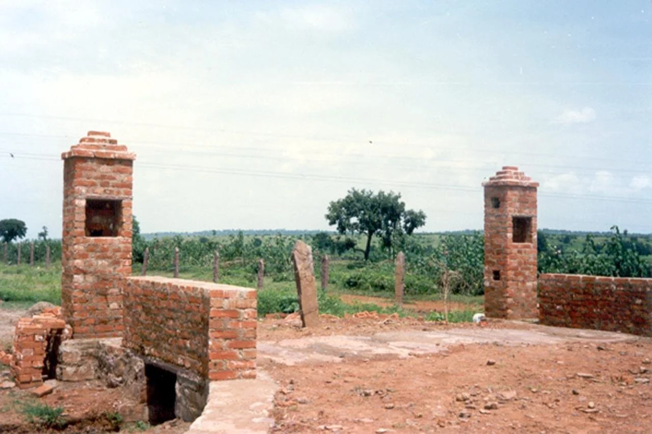 Two brick chimneys and a low brick wall on dirt ground in a rural landscape with green fields and a tree in the background under a partly cloudy sky.