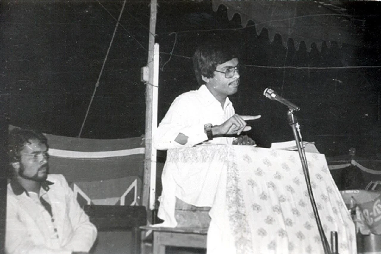 A man with glasses speaking at a podium covered with a floral cloth, gesturing with his hands, while another man sits nearby.