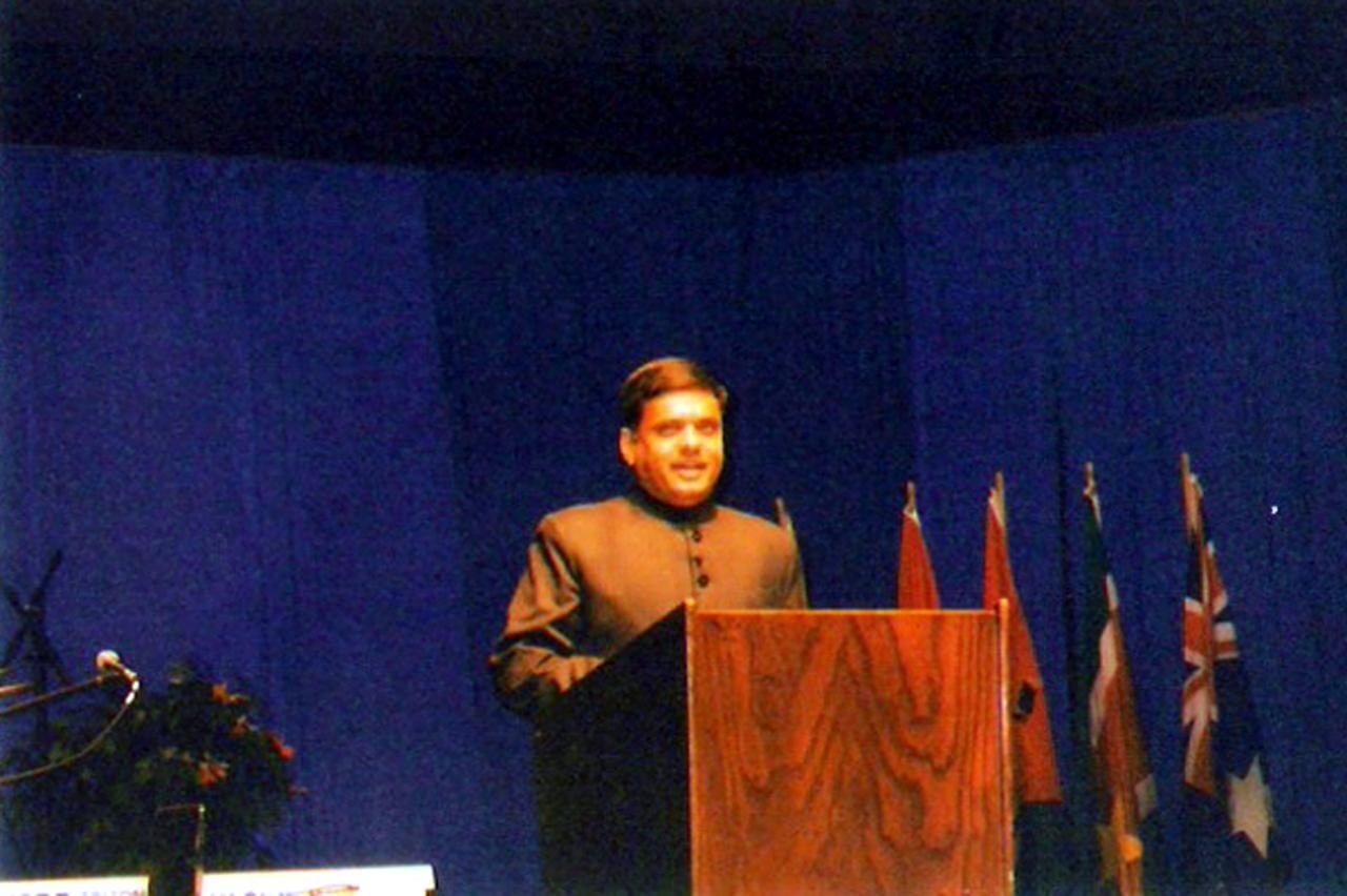 A man standing behind a wooden podium giving a speech with flags in the background and a blue curtain behind him.