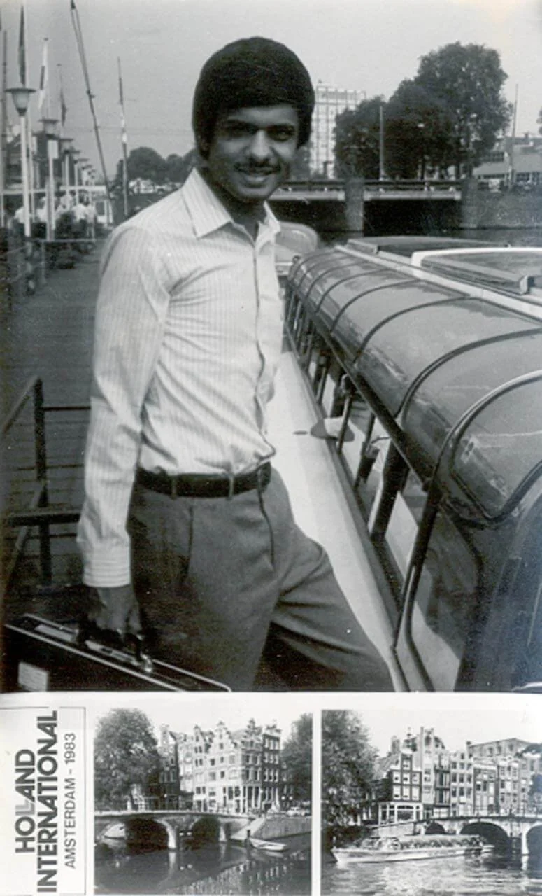 A black-and-white photo of a man with dark skin and a large afro hairstyle smiling at the camera, standing outdoors next to watercraft, with a cityscape and trees in the background.