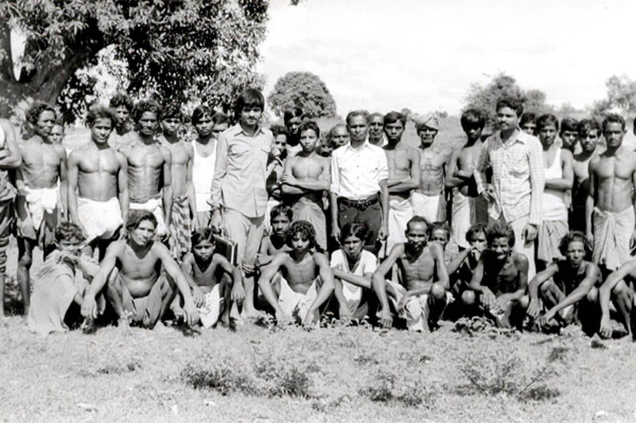 A black and white photo of a large group of men, many shirtless and wearing traditional loincloths, outdoors on a grassy field with trees in the background.