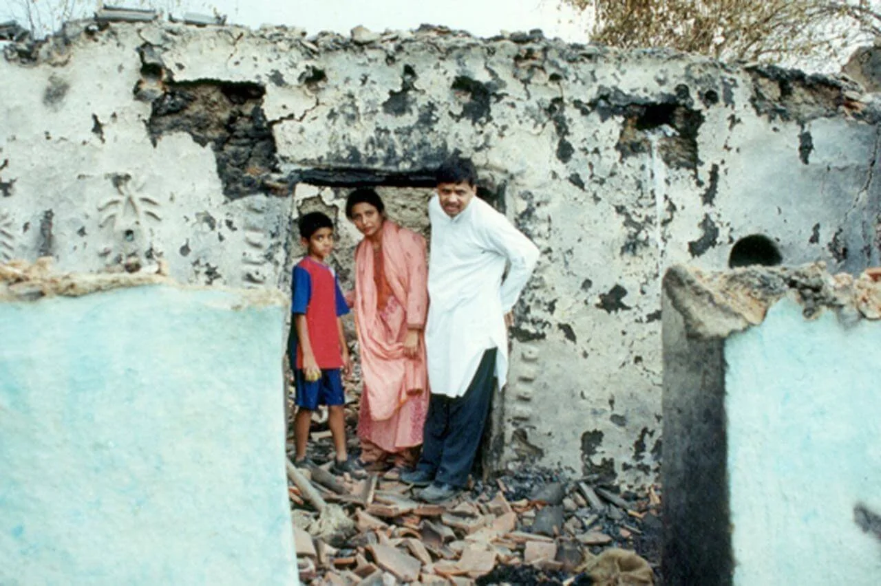 Three people, two children and an adult, standing inside a destroyed building with crumbled walls and rubble on the ground.