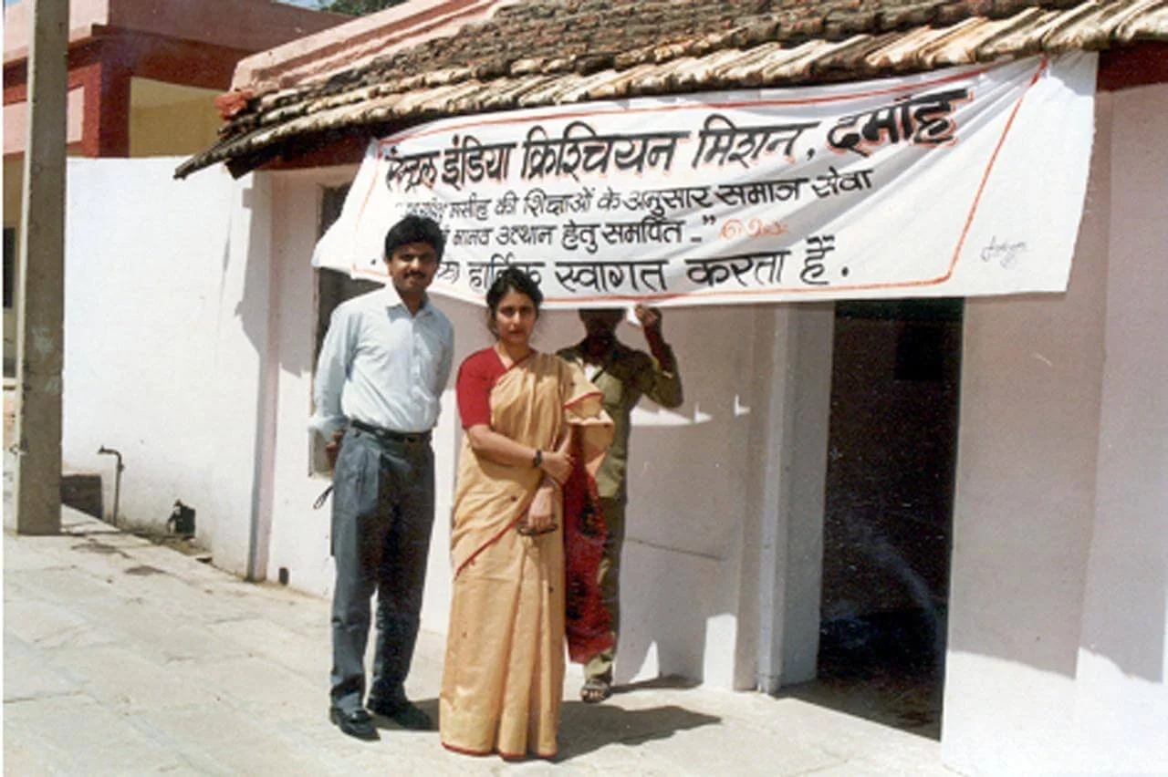 Three people standing next to a building with a banner in Hindi. The banner appears to be for a community event or celebration, with one person holding onto the banner pole.
