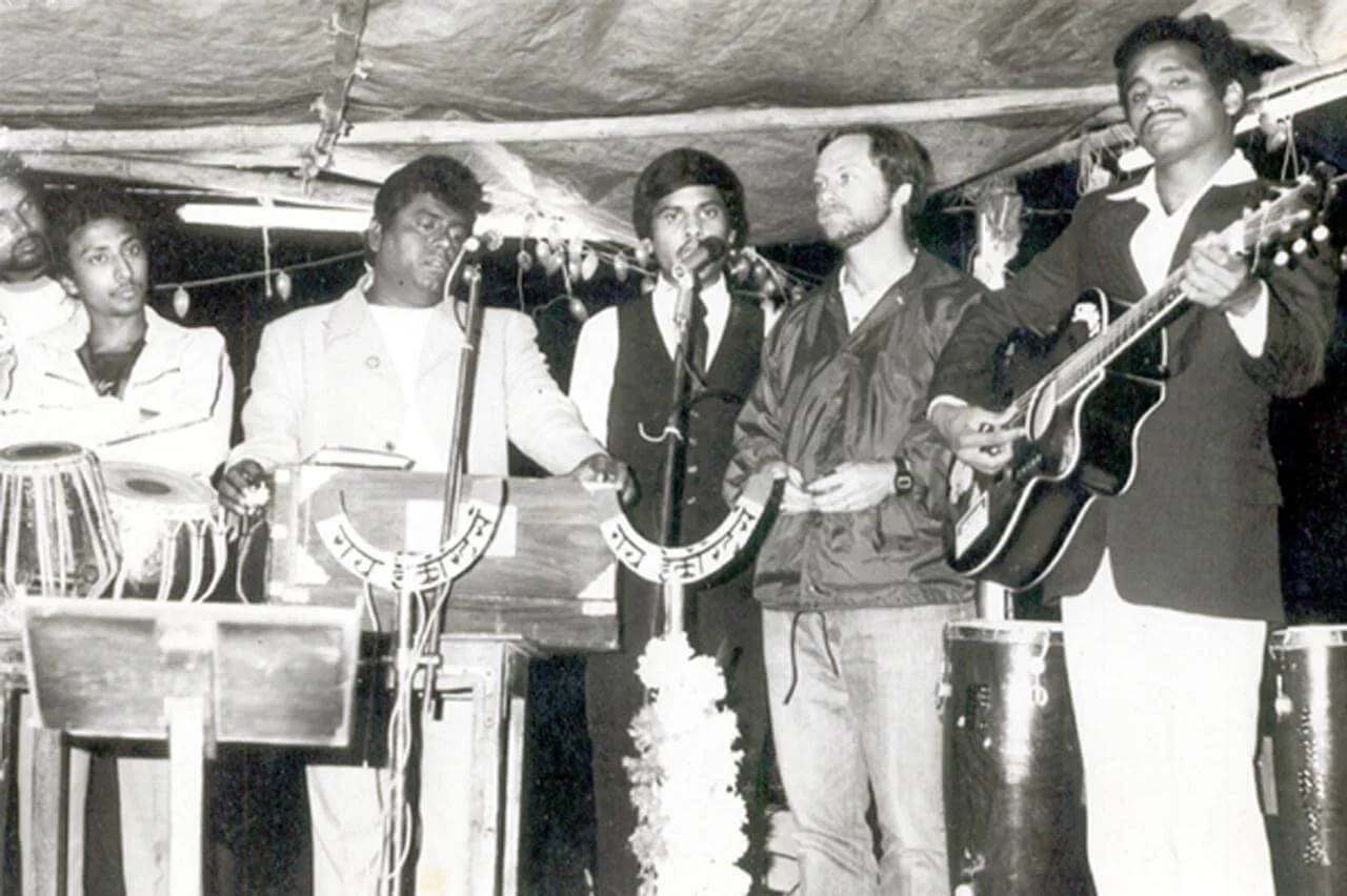 Black and white photo of five men performing together on stage with musical instruments, including a guitar and percussion, at a cultural event or celebration.