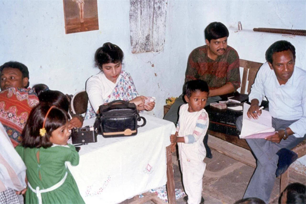 A group of children and adults sitting around a table and on benches in a room, with some children talking and looking at the camera, and a woman holding money.