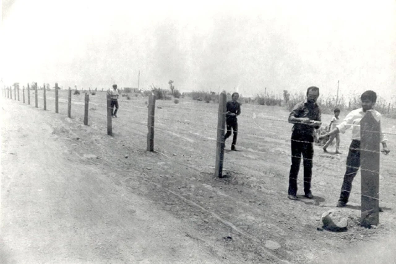 Children playing near a barbed wire fence on a dirt field in a rural area.