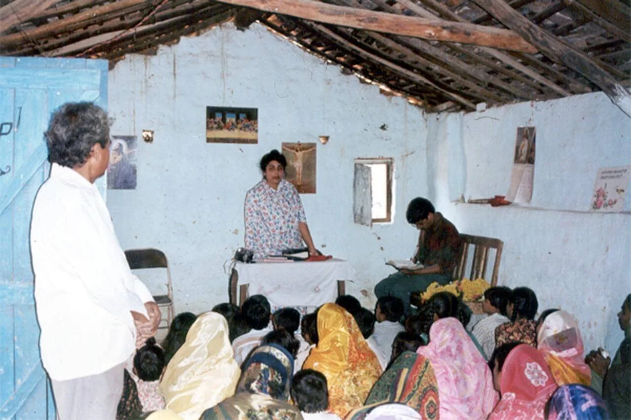 A gathering inside a rustic room with white walls and a wooden ceiling, where a woman is speaking at a table in front of a seated audience of men, women, and children. One man stands to the side, and another woman is reading or writing. The audience 
