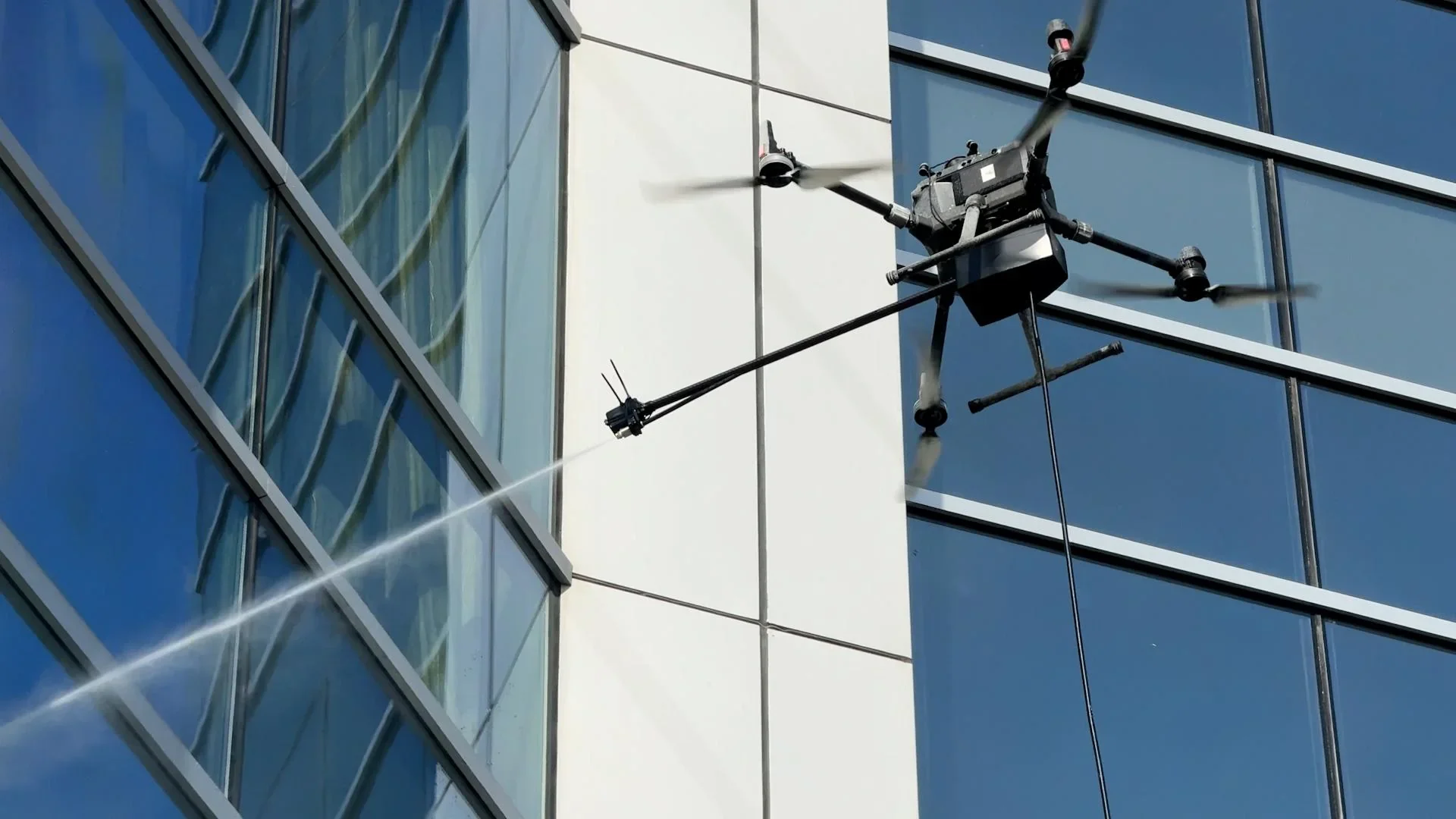 A drone equipped with a spray nozzle is cleaning or disinfecting the exterior windows of a modern glass office building.