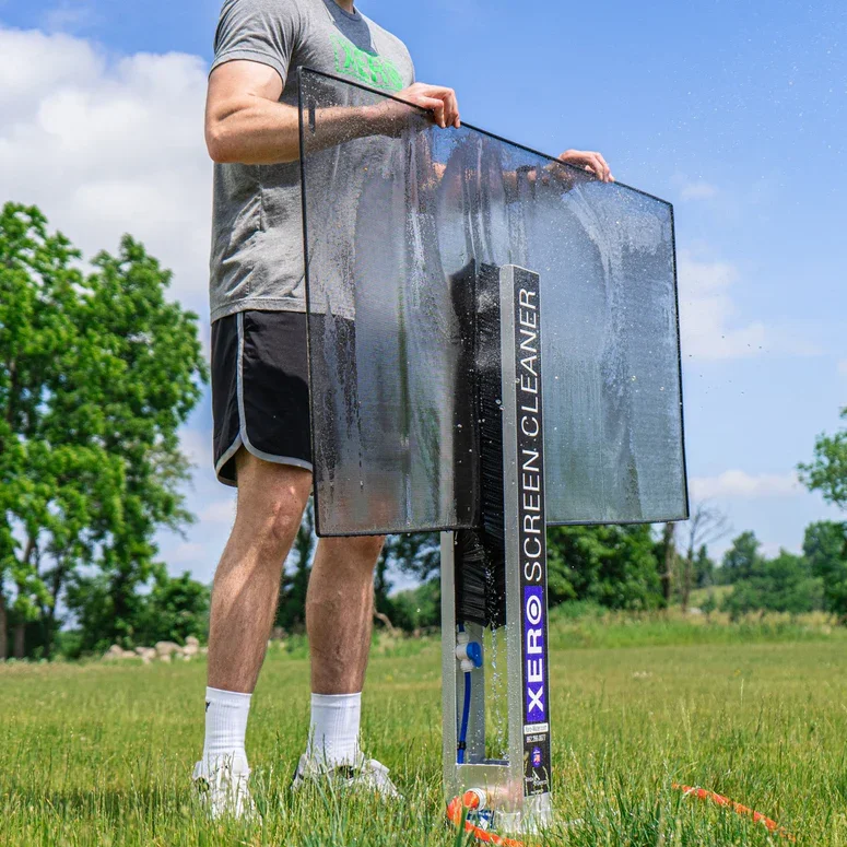 A person in a gray t-shirt and black shorts is setting up a large outdoor water screen with a water spray mechanism, on a grassy field with trees in the background.