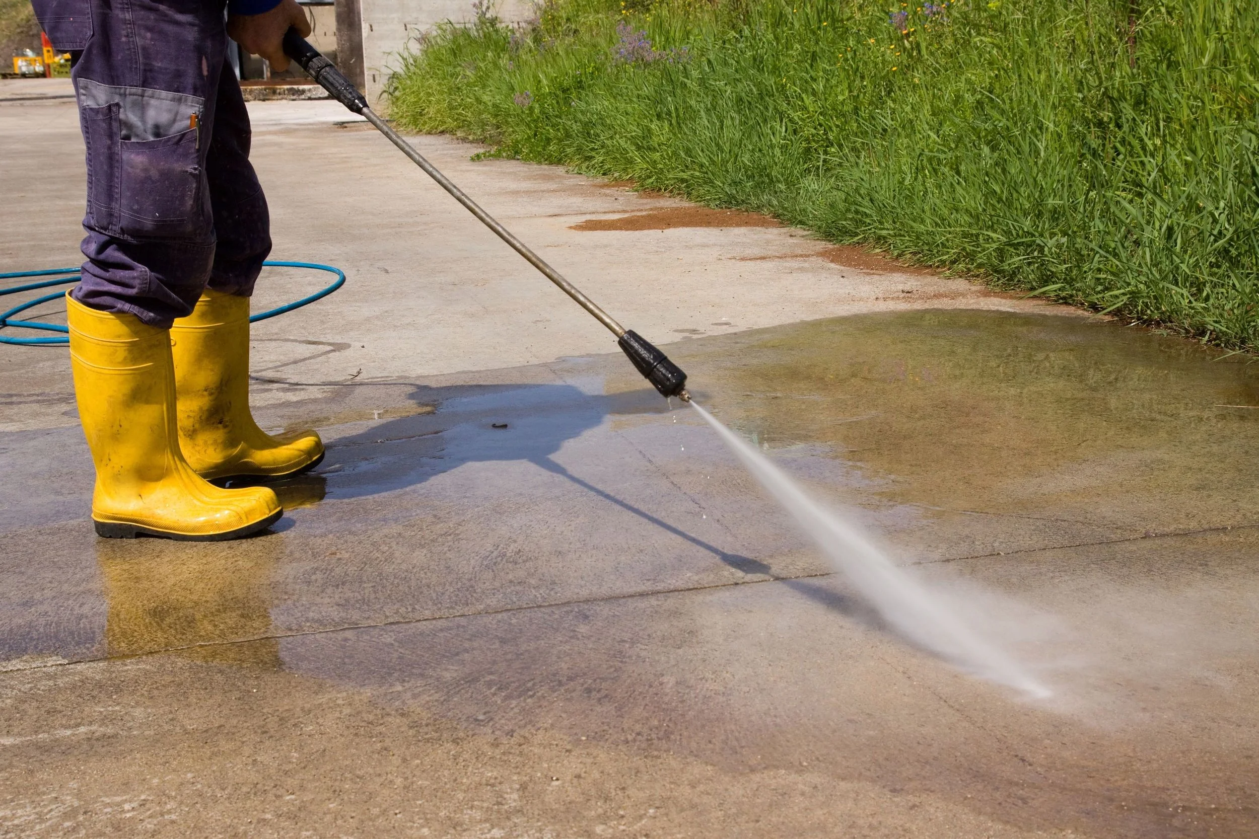 A person wearing yellow rubber boots and navy pants is pressure washing a concrete sidewalk near grass and purple flowers.