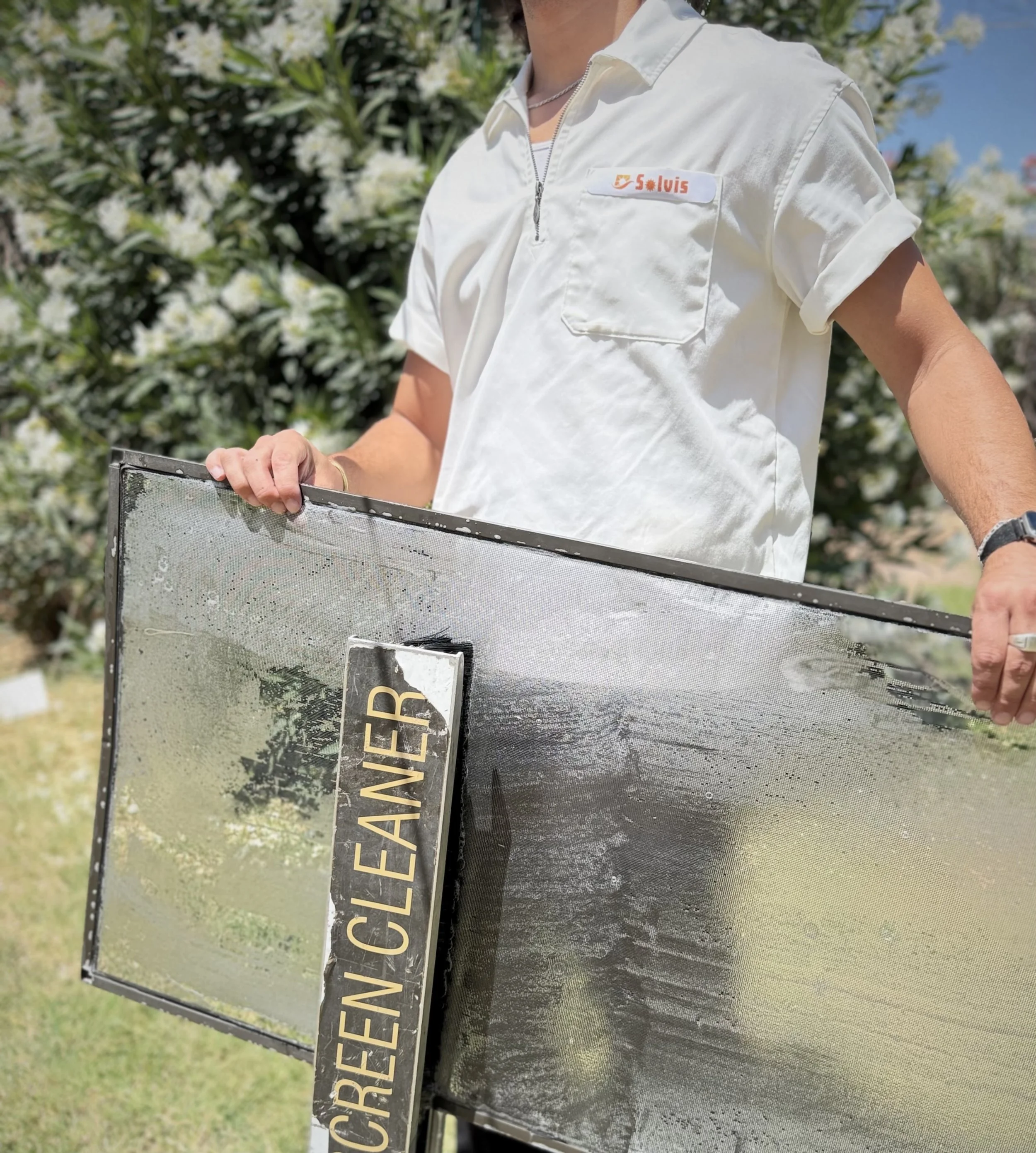 A person in a gray t-shirt and black shorts is setting up a large outdoor water screen with a water spray mechanism, on a grassy field with trees in the background.