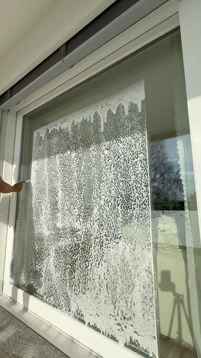 A sliding glass door with a frost or snow pattern on the glass, partially opened, with a reflection of trees and sky outside.