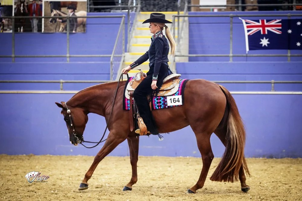 A woman in western attire riding a chestnut quarter horse in an indoor arena with Australian flag in the background. Showing horse at the lope gait.