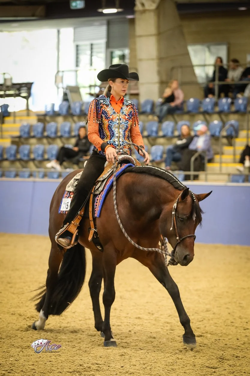 A cowgirl wearing a black cowboy hat, an orange shirt, and a colorful western jacket riding a dark brown quarter horse inside an indoor arena in Sydney Australia showing in western pleasure futurity class loping horse.