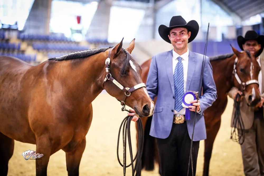 Man in a light blue blazer, striped tie and cowboy hat standing with a brown quarter horse, holding a ribbon, at an indoor western performance show in Australia.
