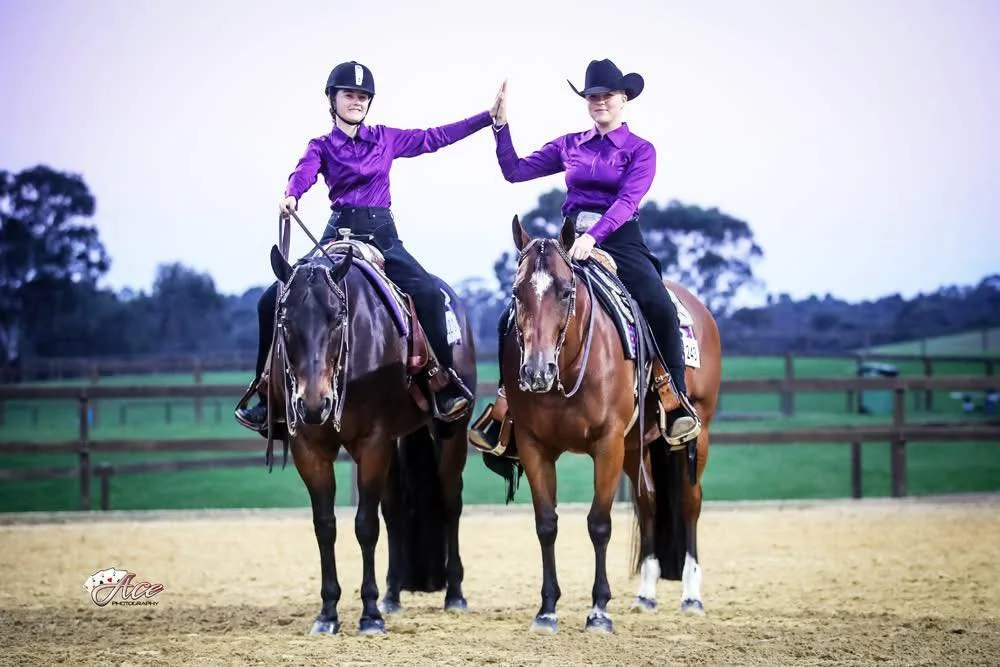 Two women riding horses in an outdoor riding arena, wearing matching purple shirts and black helmets, giving each other a high five.
