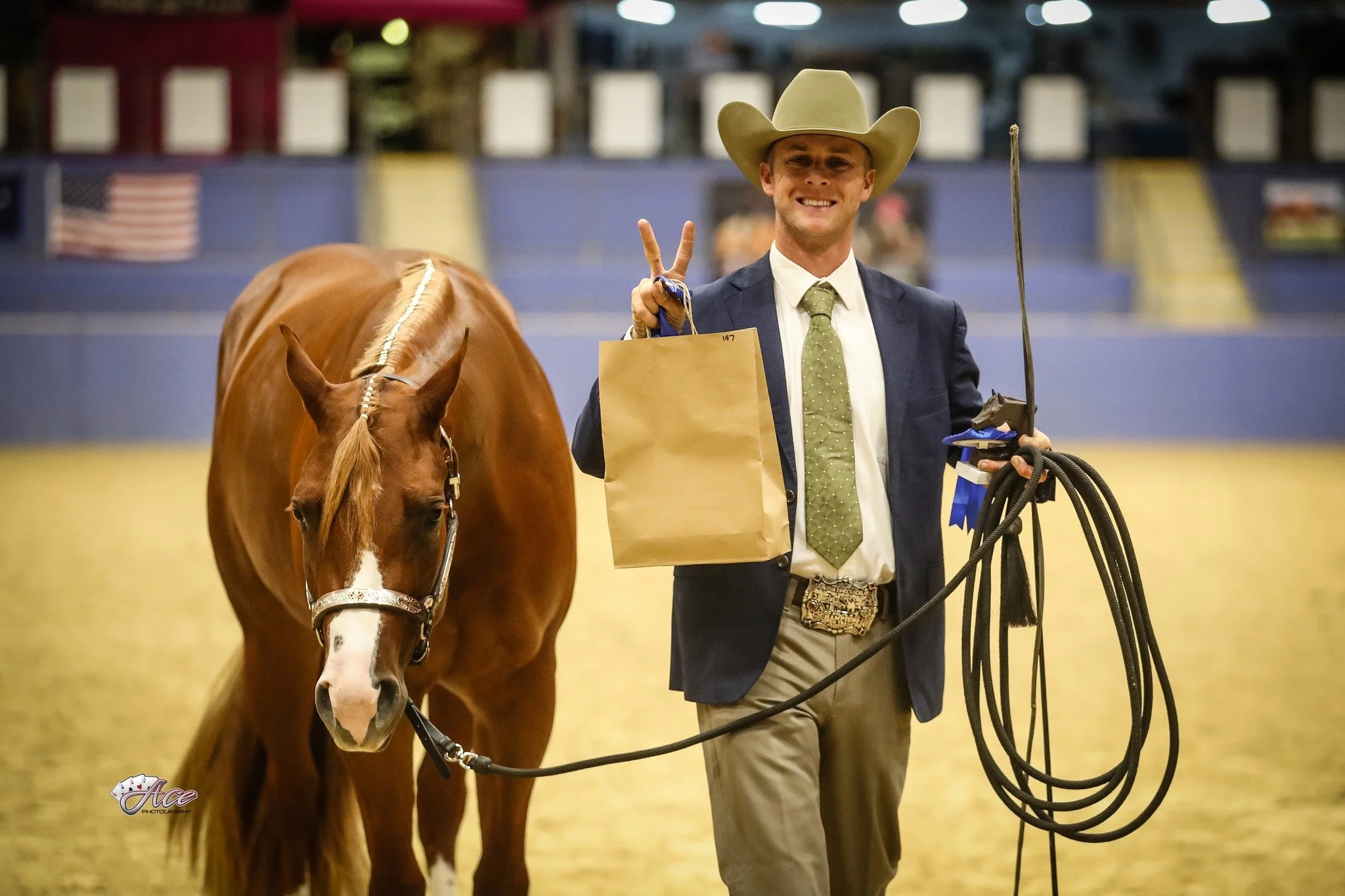 A man wearing a cowboy hat and formal attire holding a horse's reins with a ribbon on the horse's bridle, smiling and making a peace sign with his hand, winning lunge line event western performance show.