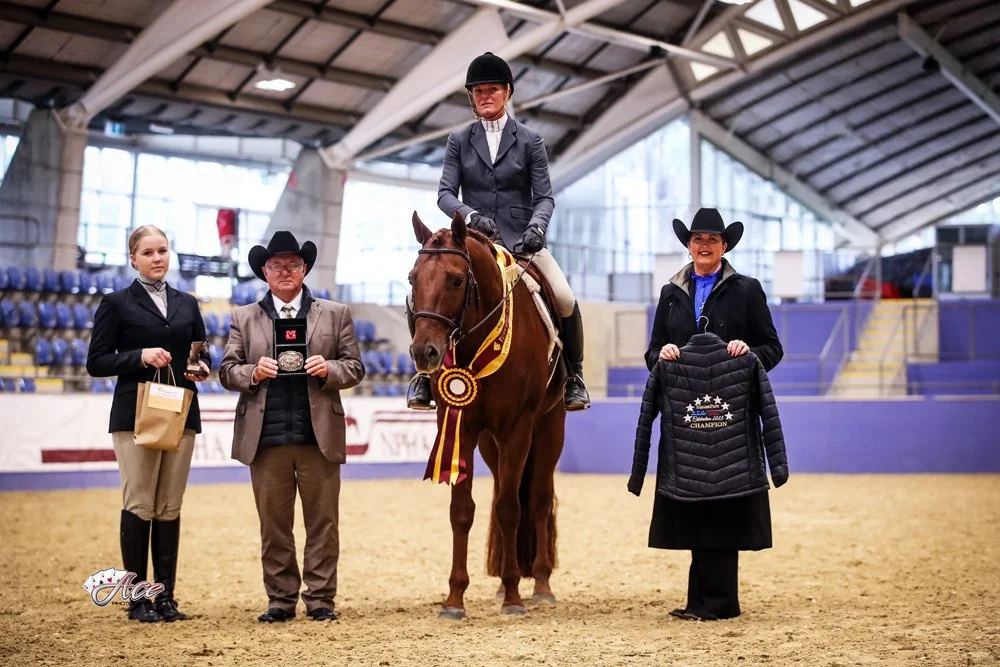 A woman on a hunter under saddle horse receiving a ribbon at a western performance competition, flanked by three people, inside an indoor arena at SIEC.