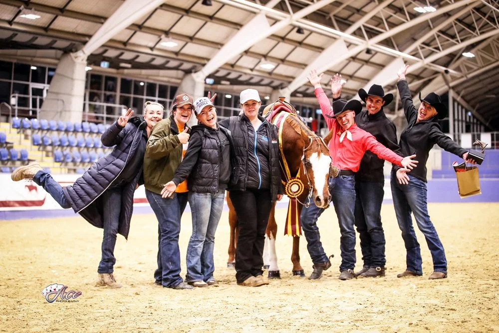 Group of seven people smiling and celebrating with a horse in an indoor arena. Some are wearing cowboy hats and casual clothing at Sydney International Equestrian Centre.