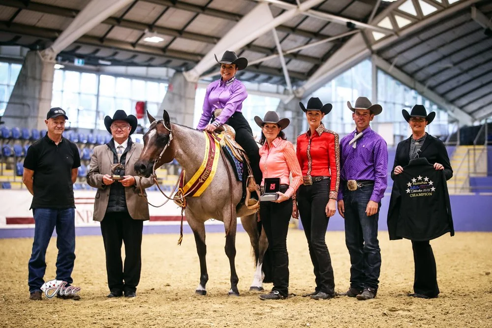 Group of six people, including a woman on a horse, celebrating a futurity championship. The woman is wearing a purple shirt and black cowboy hat. The others are in cowboy hats and Western attire, some holding awards and a champion jacket.
