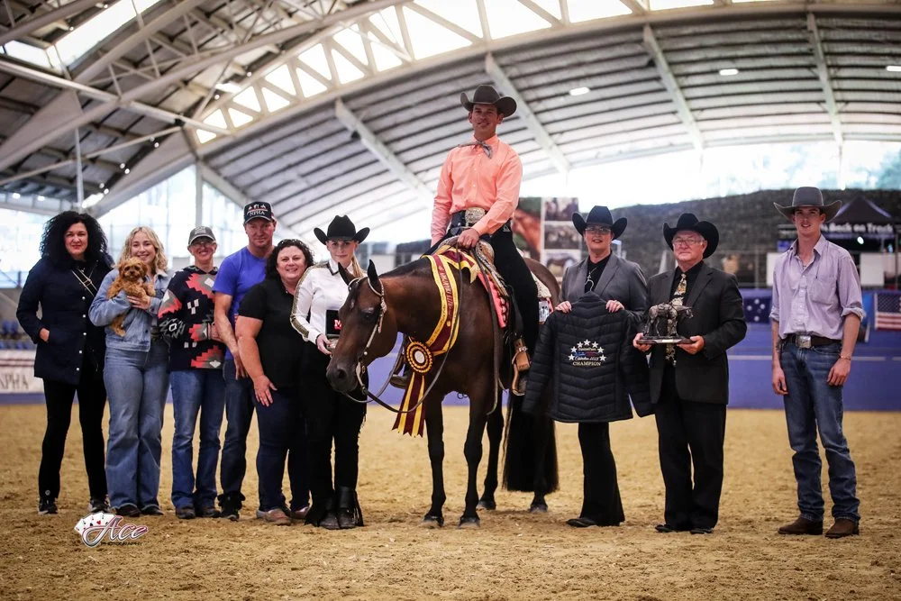 A young man in cowboy attire sits atop a horse with ribbons, surrounded by a group of people inside an indoor SIEC. The group includes men and women, some dressed in formal attire and cowboy hats, holding ribbons. Winning Western Pleasure Futurity.
