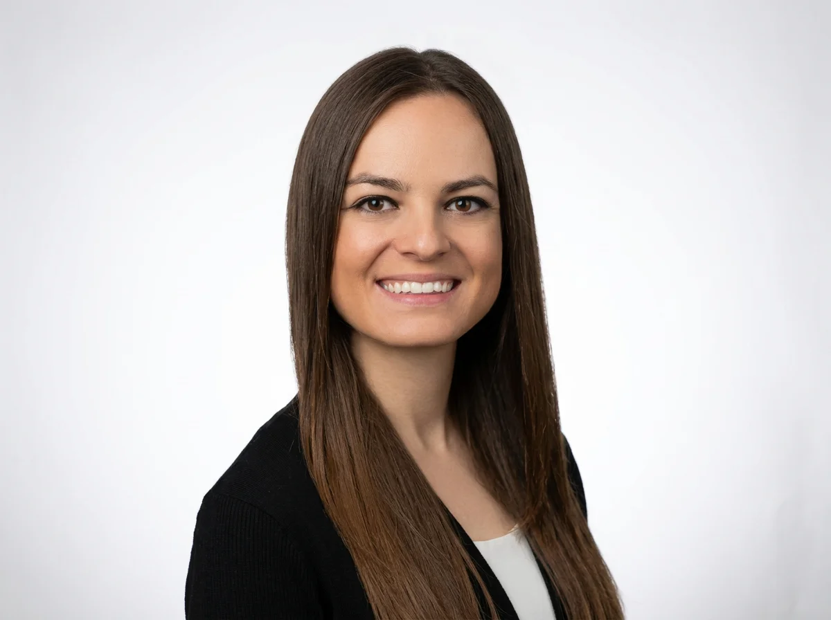 A woman with long brown hair smiling at the camera, wearing a black blazer and white top, against a plain white background.