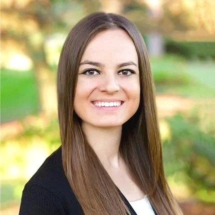 Portrait of a smiling young woman with long brown hair, outdoors in a park with green trees and autumn leaves in the background.