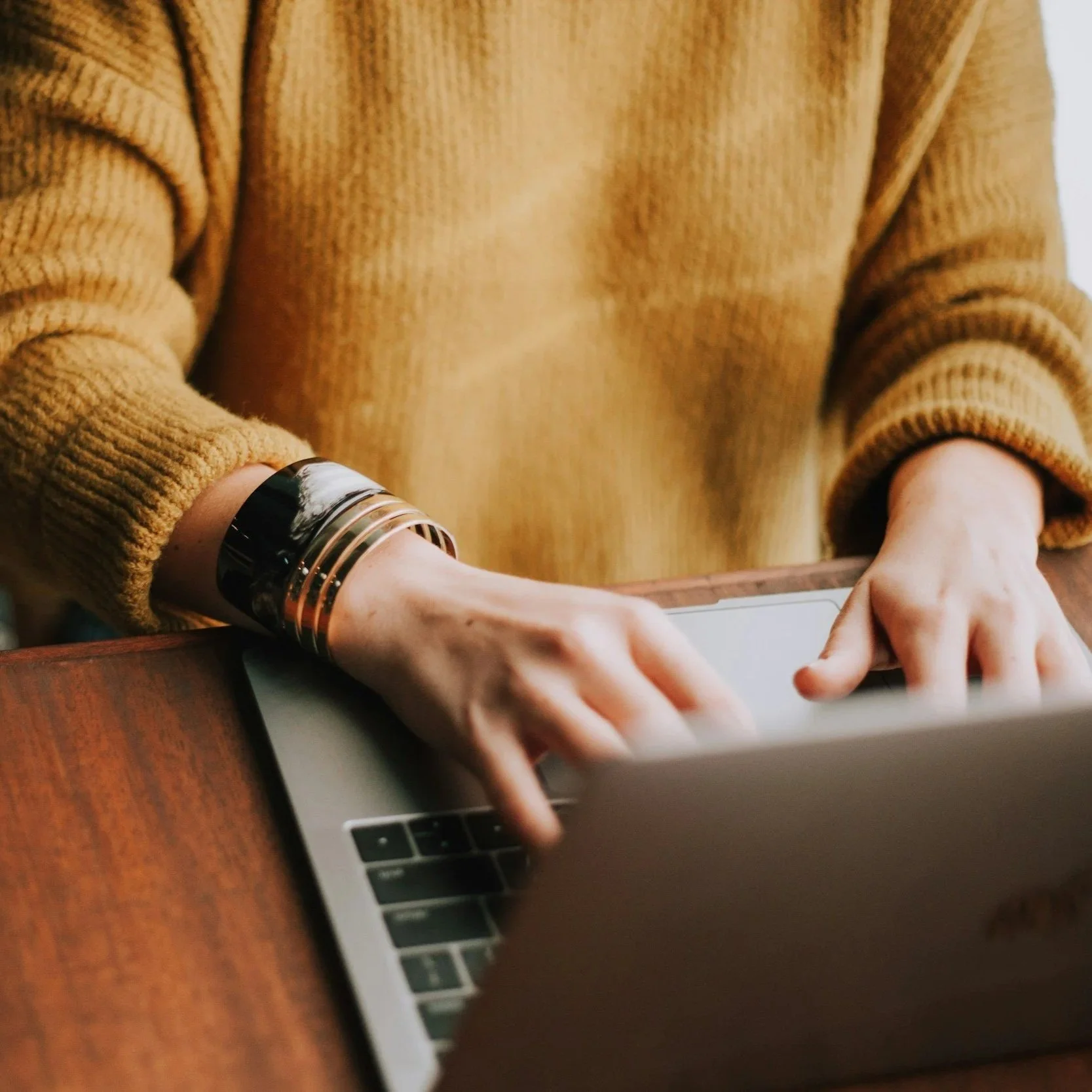 Person wearing a yellow sweater and large metallic bracelet, working on a laptop at a wooden table.