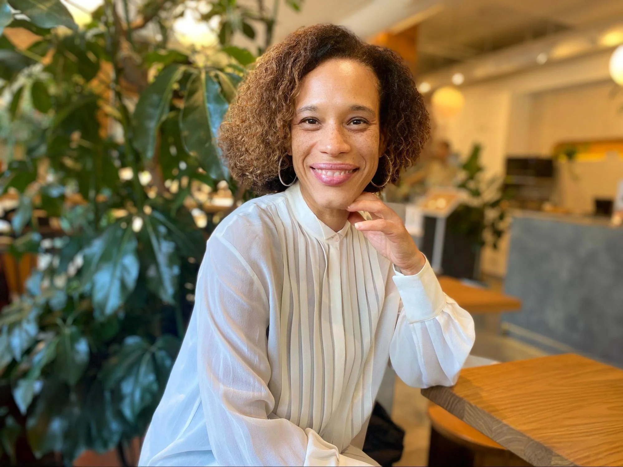 A woman with curly hair and hoop earrings smiling while sitting at a wooden table in a warmly lit cafe with large green plants in the background.