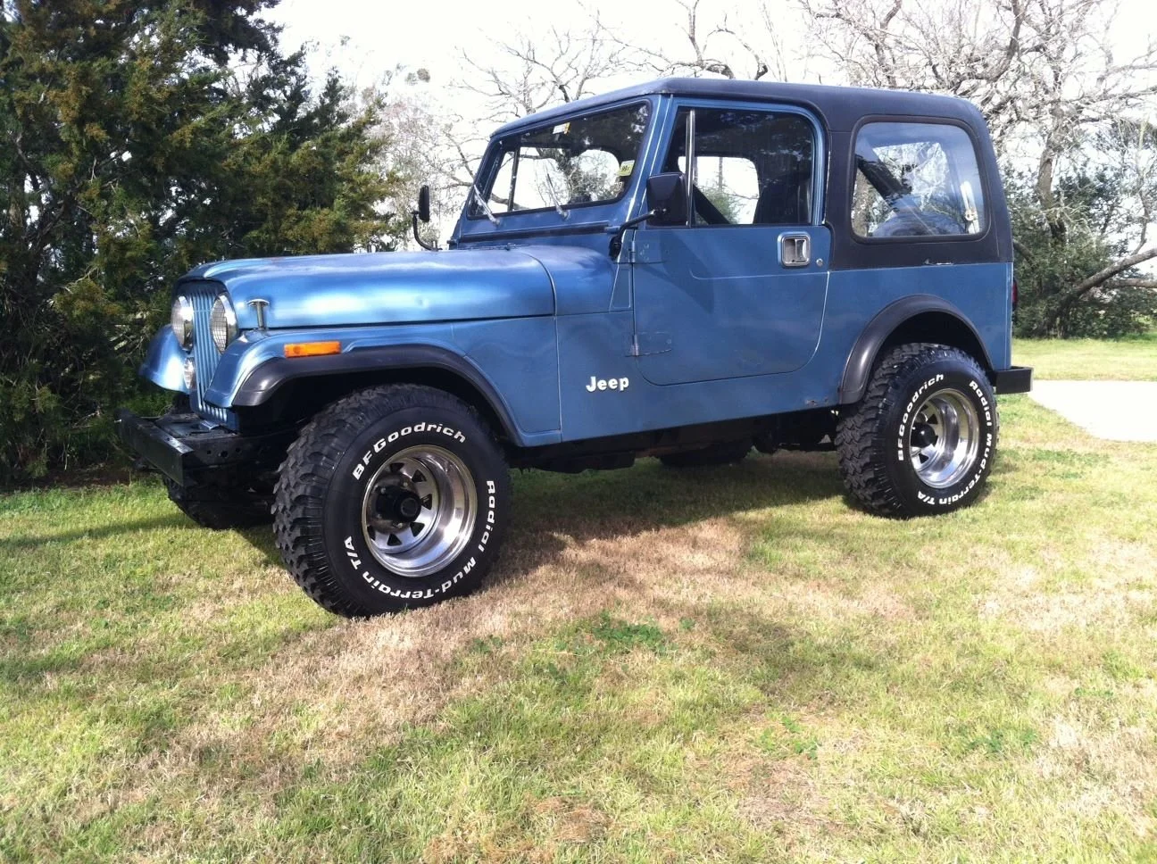 Blue vintage Jeep with large off-road tires parked on grass near bushes and trees.