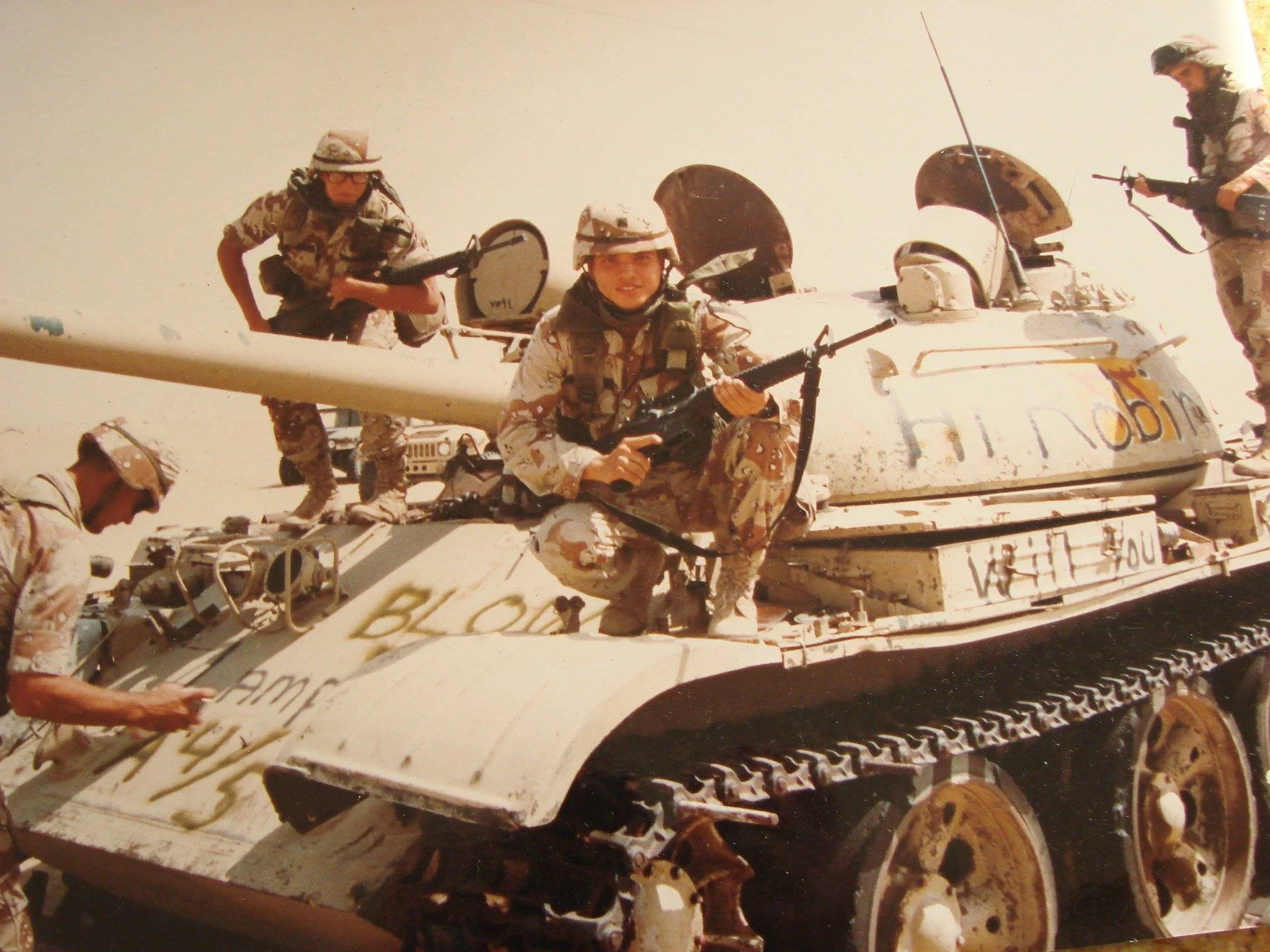 Soldiers in desert camouflage gear sitting and standing on a military tank, some holding rifles, in a desert environment.