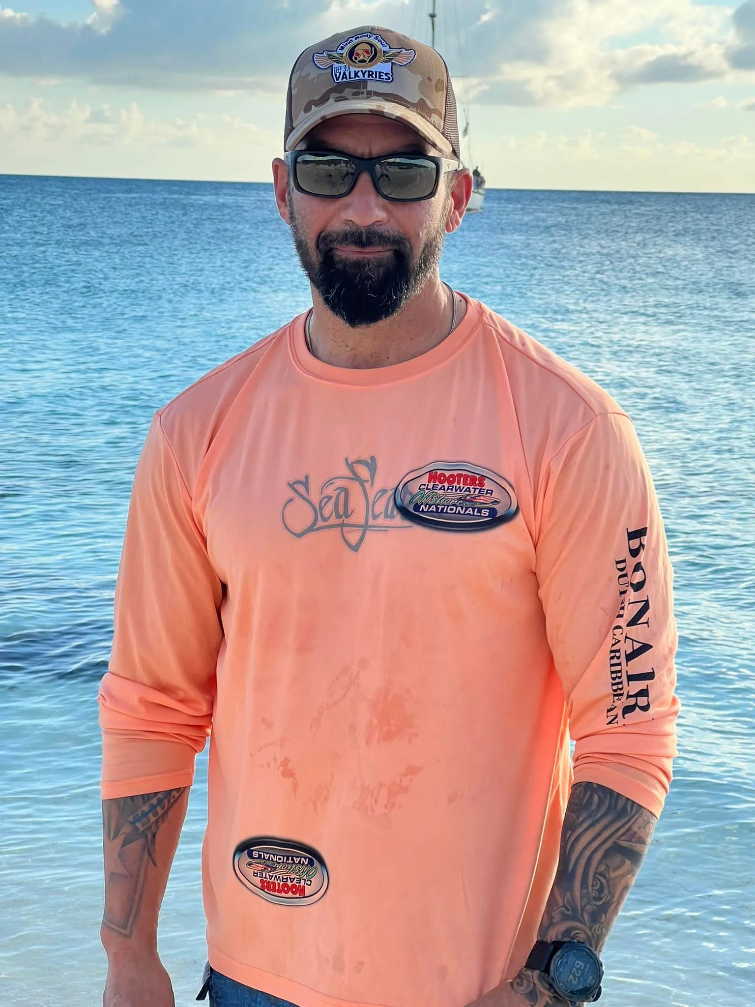 A man wearing sunglasses, a beige camouflage hat with a Valkyries logo, and a peach long-sleeve shirt with various patches, standing in front of the ocean with a boat in the distance.