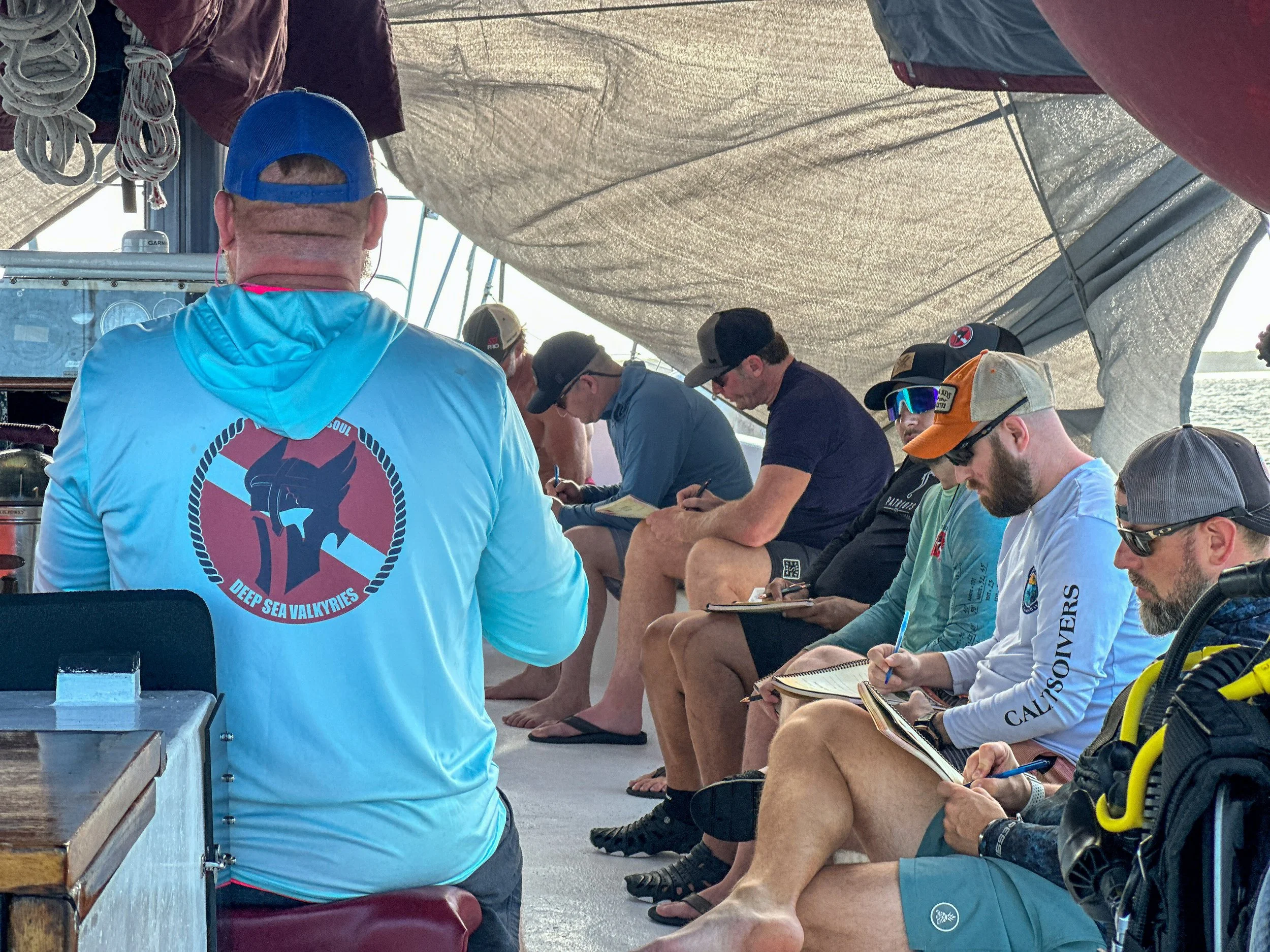 group of men on boat deck writing in workbooks during counseling session. Counselor is wearing a shirt with Deep Sea Valkyries logo