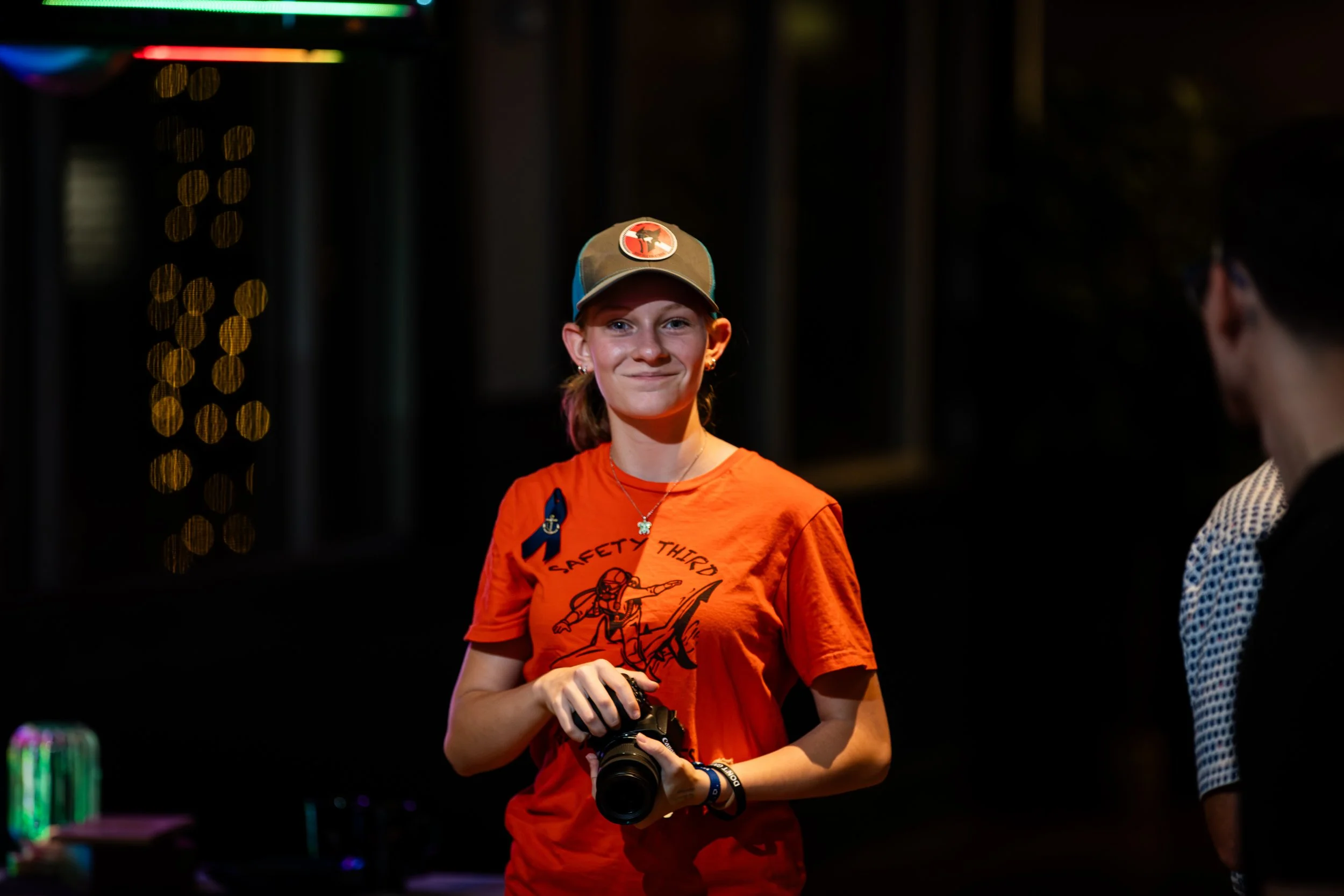 A young woman with a camera around her neck and a safety third t-shirt, smiling, standing in a dimly lit room with colorful bokeh lights in the background, and another person partially visible on the right.