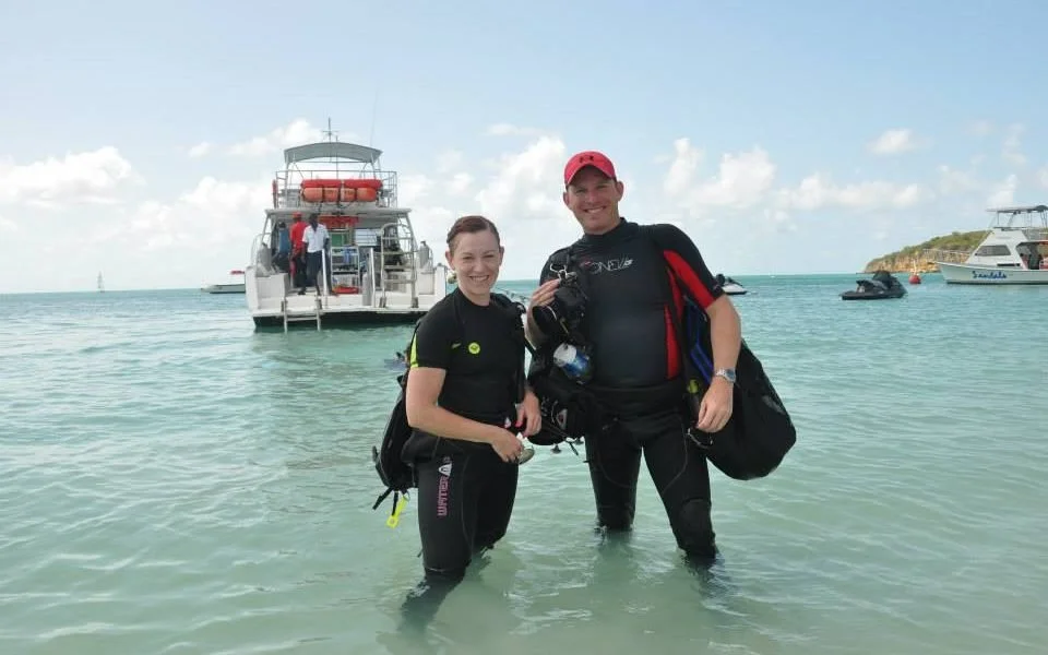 Two scuba divers standing in shallow water near boats, with a clear sky and small island in the background.