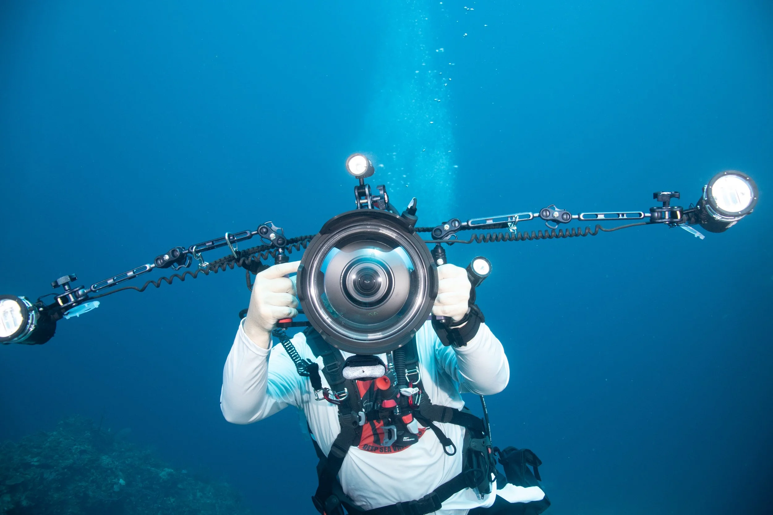 Underwater photographer taking a picture with a specialized camera with lights, wearing diving gear including a wetsuit and harness, surrounded by blue ocean water.