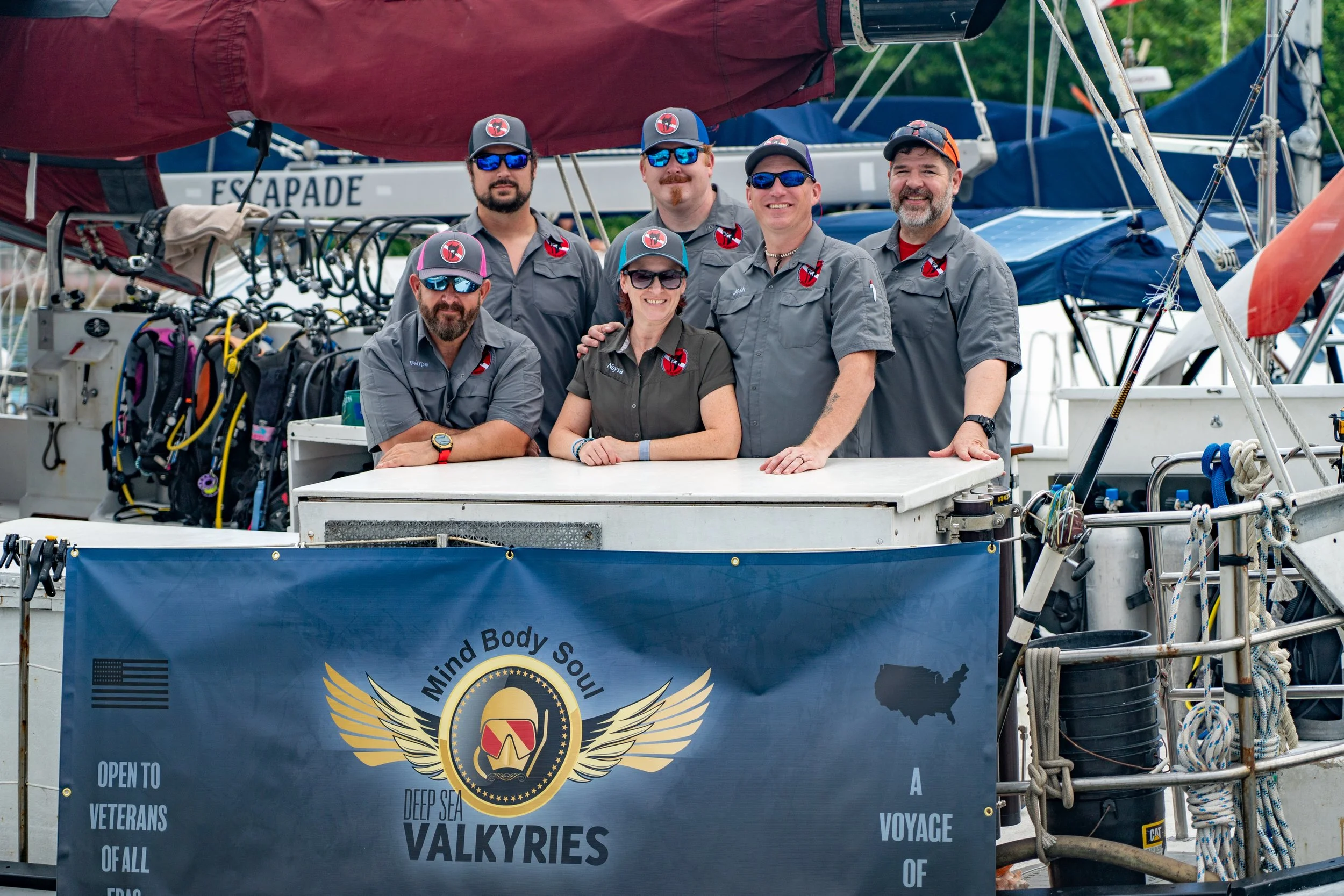 Group of six people, five men and one woman, wearing matching gray shirts with logos, posing together on a boat with a banner that reads "Mind Body Soul" and features an American flag silhouette.