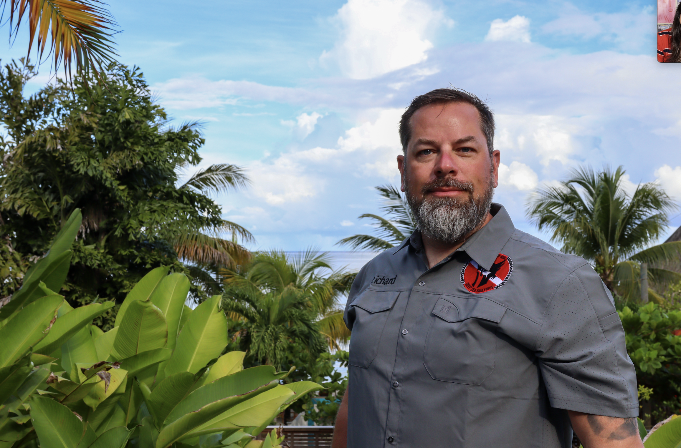 A man with a beard wearing a gray shirt standing outdoors among lush green tropical plants and palm trees, with a partly cloudy sky in the background.