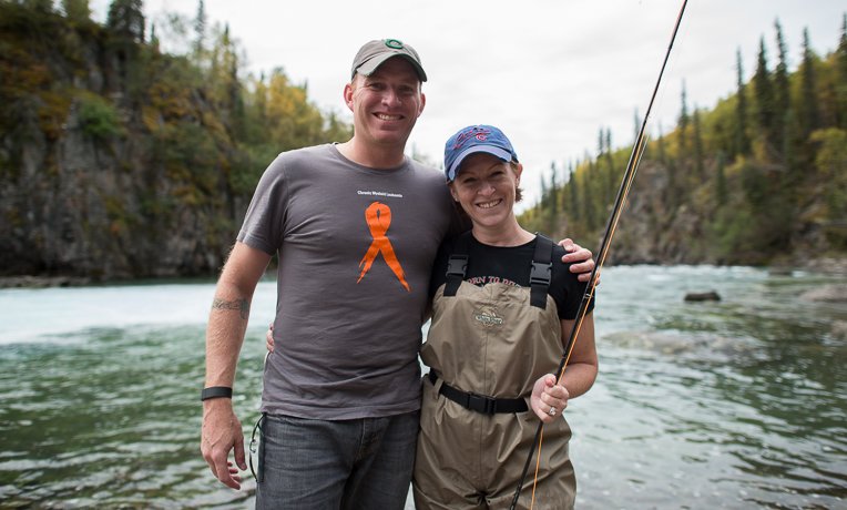 A man and woman standing together by a river, smiling, with trees and rocky cliffs in the background. The woman is holding a fishing rod, and they are dressed casually for outdoor activity.