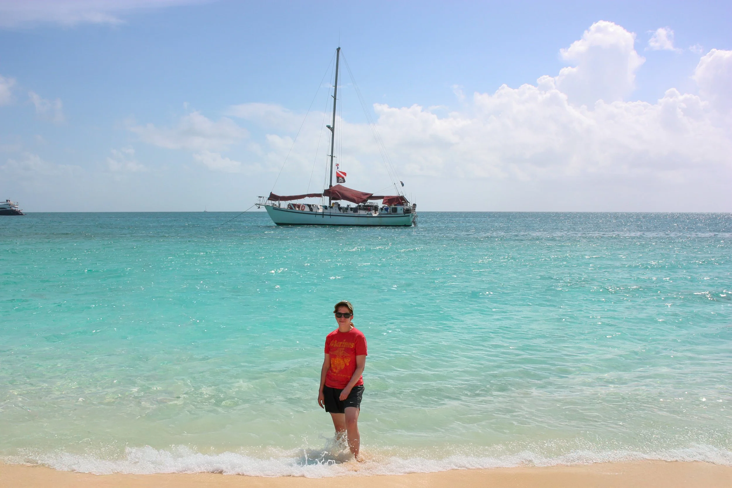 A person in red shirt and black shorts standing in shallow clear turquoise water at a sandy beach with a sailboat anchored in the background under a partly cloudy sky.