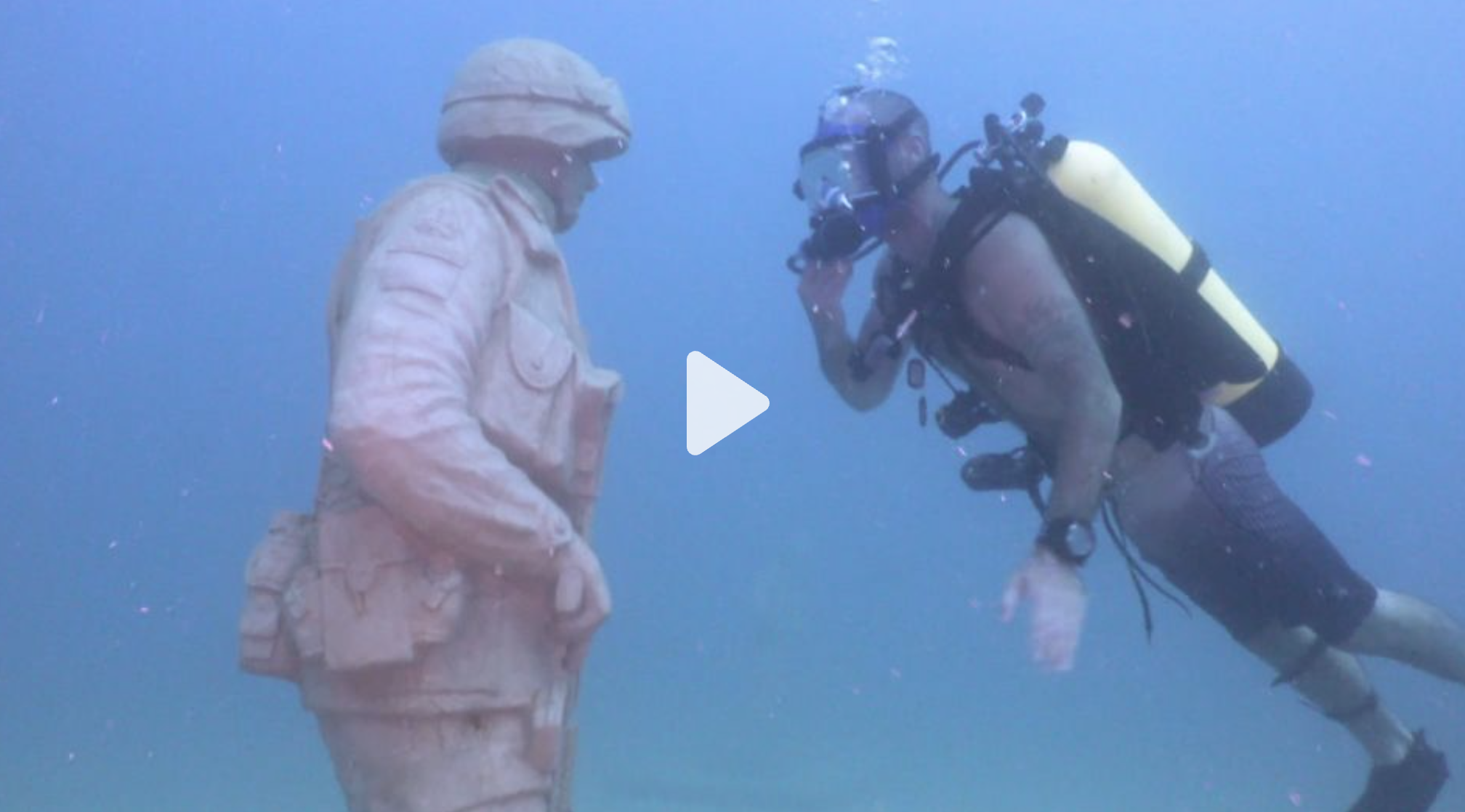 A diver in a scuba suit and mask appears to be communicating with a mannequin dressed as a lifeguard underwater.