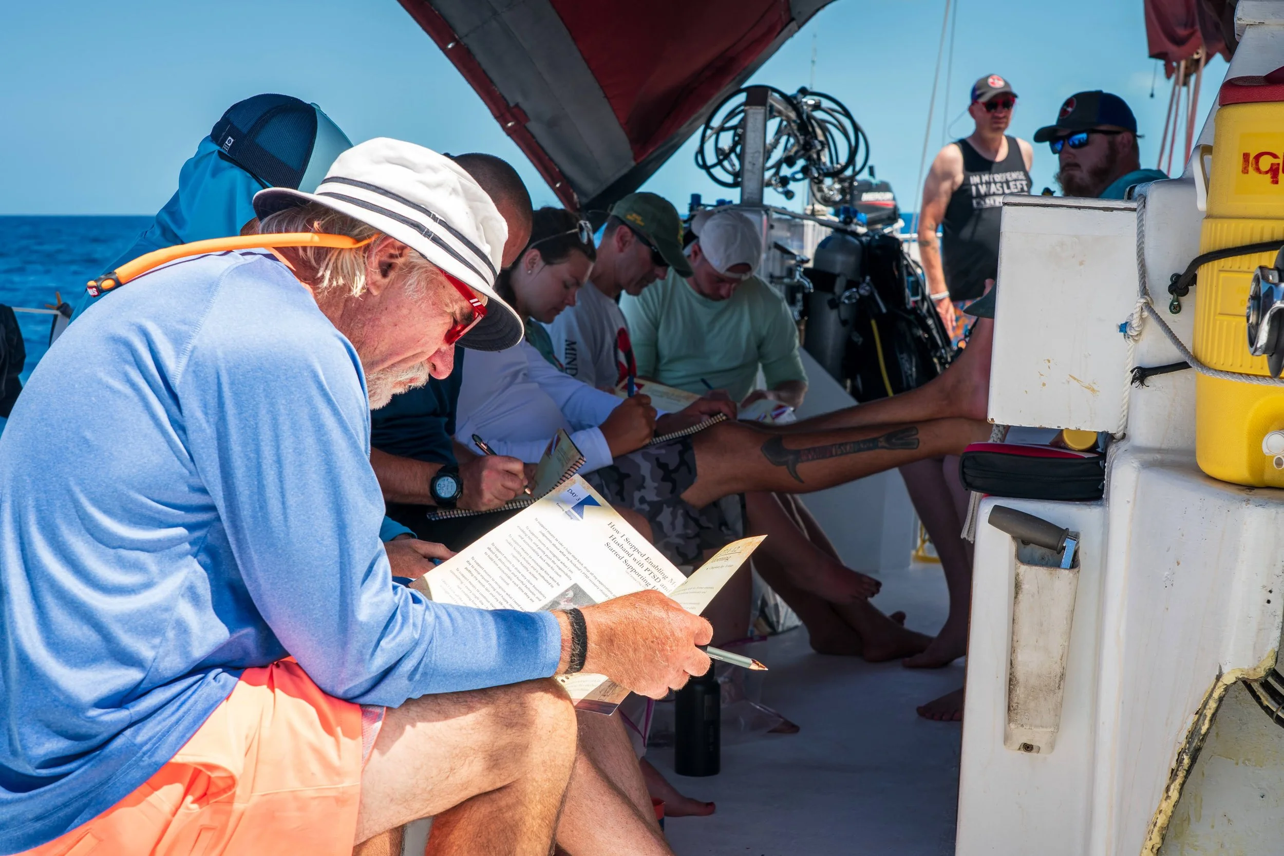 A group of people sitting on a boat, writing in notebooks, with the ocean in the background. One person in the foreground is reading a pamphlet.