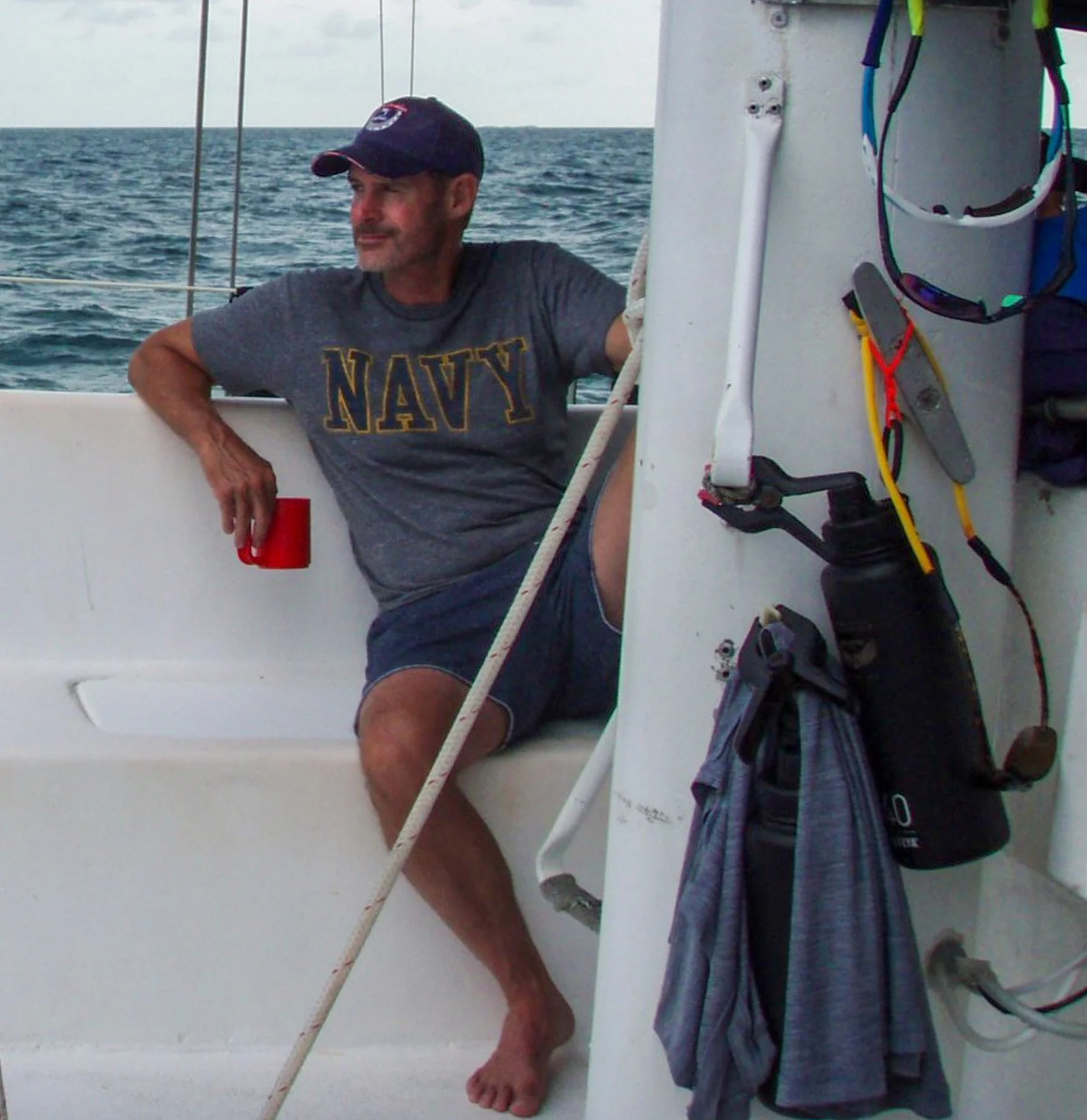 Middle-aged man sitting on a boat, holding a red cup, wearing a navy T-shirt, shorts, and a cap. The boat is on the water with a gray sky above.