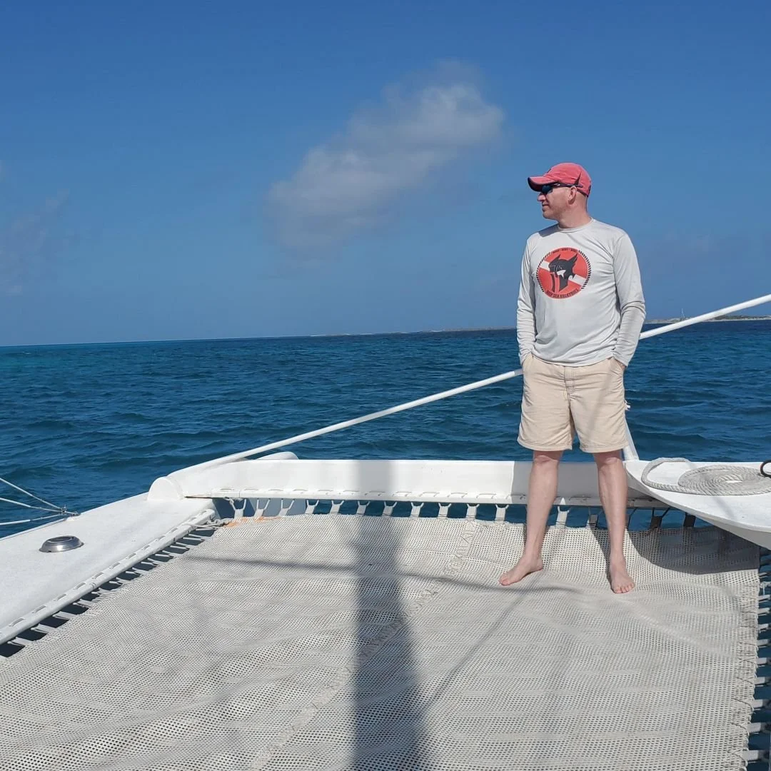 A man standing barefoot on a boat deck, wearing a gray long-sleeve shirt with a graphic, beige shorts, a red cap, and sunglasses, looking out at the ocean under a partly cloudy sky.