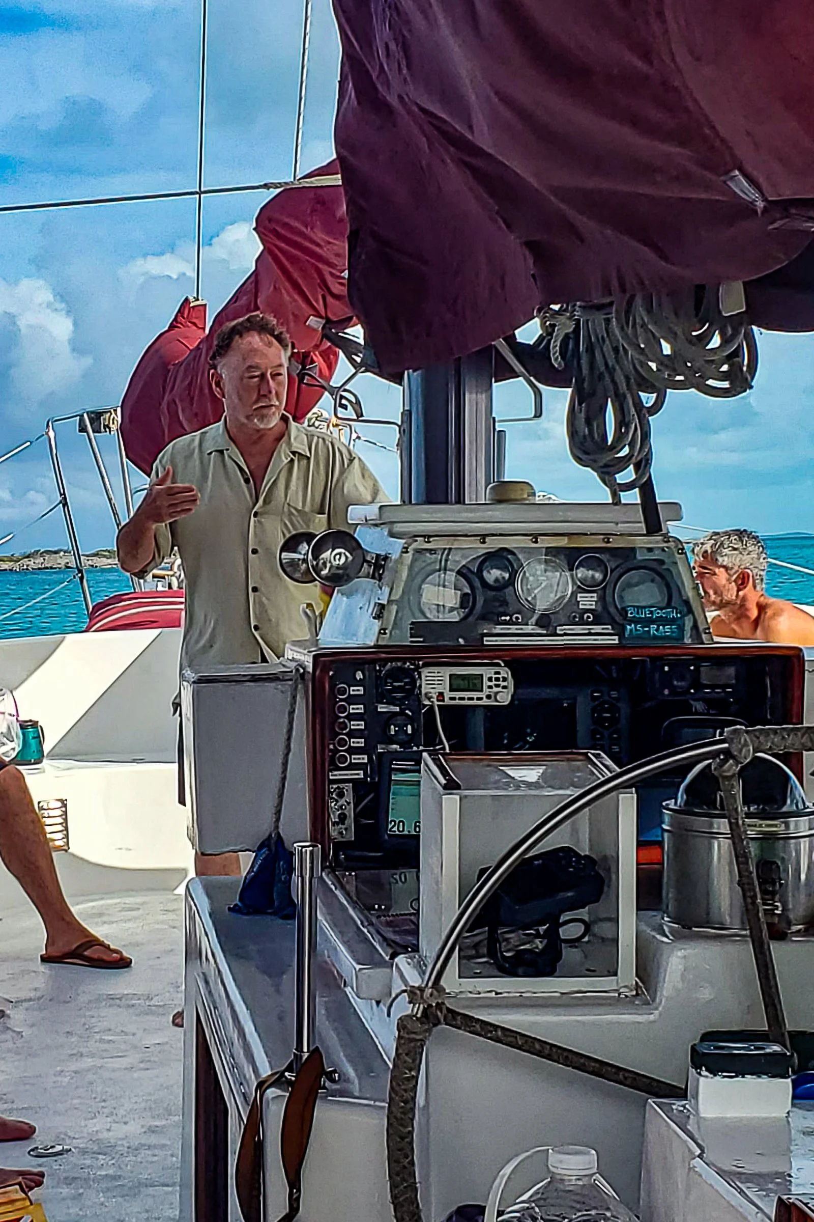 A man is speaking and gesturing with his hand on a boat deck, surrounded by equipment, with other people and the ocean in the background.